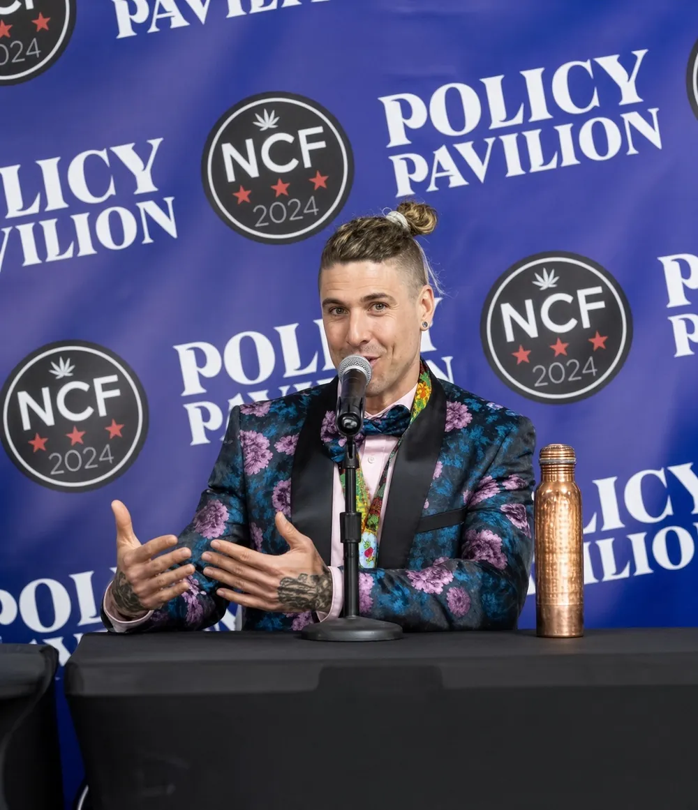 Levi Lyon, in a floral jacket, speaking into a microphone at a table in front of a Policy Pavilion, NCF 2024 backdrop.